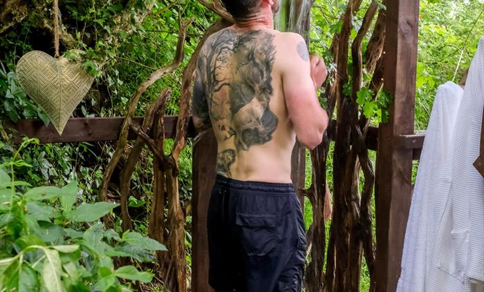 A man stands in a outdoor shower surrounded by woodland.